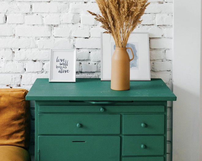 A green dresser with a frame with a saying in it and a picture behind a brown vase full of feathers.